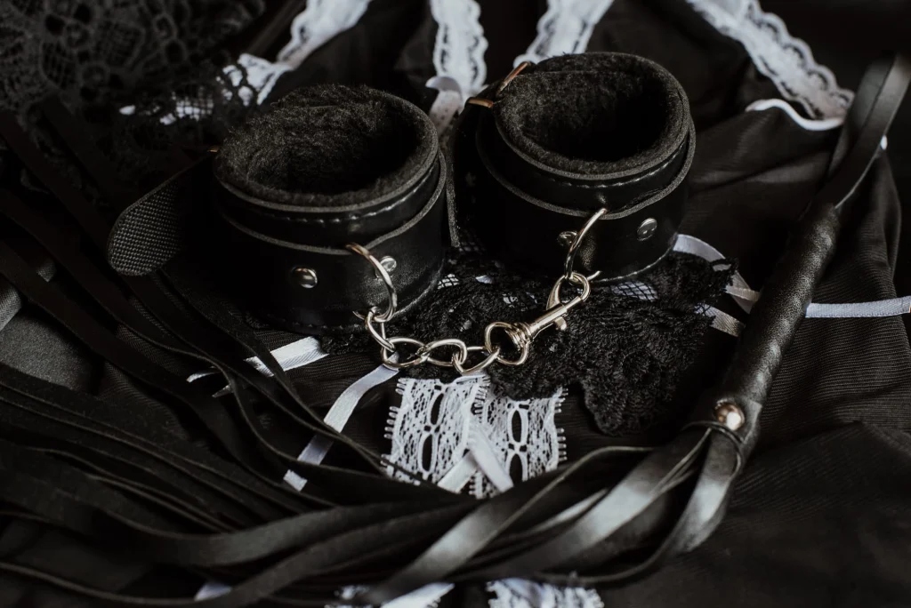 Black leather bondage cuffs joined by a chain, with a flogger and lace: restraint BDSM