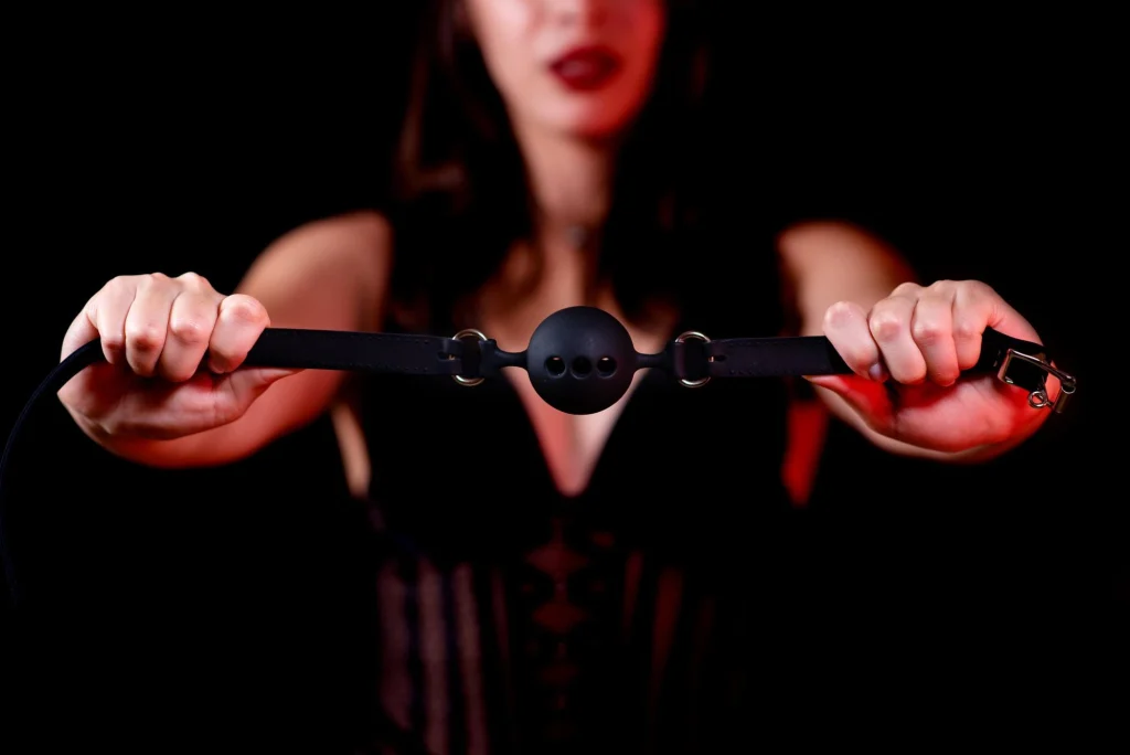 A red-lipped mistress holding a gag in low light