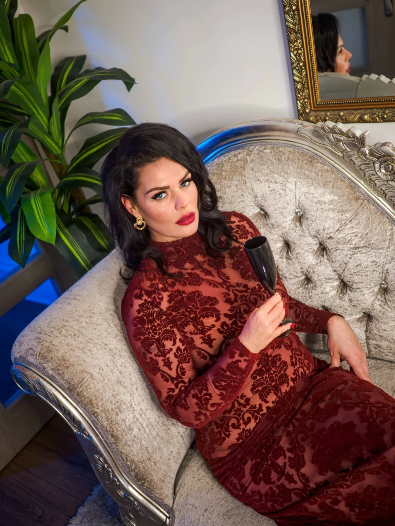 Mistress Charlotte O’Winter, London mistress, reclining on a silver chaise longue in a red lace dress