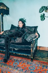 Mistress Charlotte O’Winter leaning back on a black sofa in a corset and thigh-high boots, London dominatrix