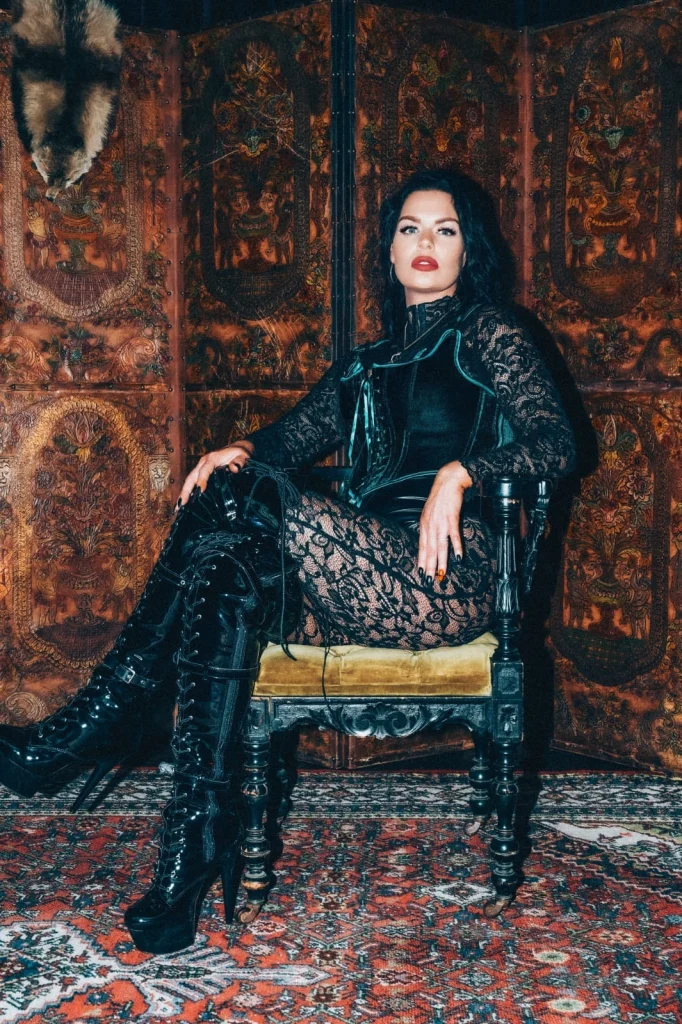 Mistress Charlotte seated before a carved leather screen in black lace and boots