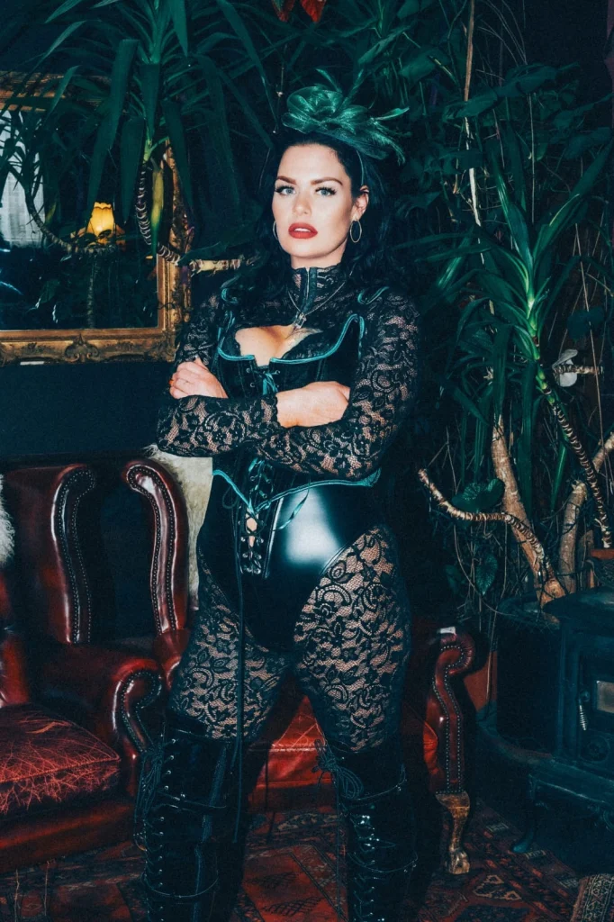 Mistress Charlotte with arms folded among palms, in a black corset and lace