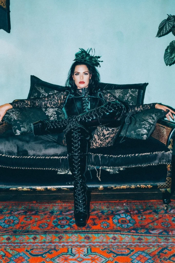 Mistress Charlotte seated with her arms outstretched along a black sofa, wearing a feathered headpiece