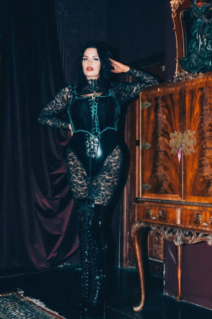 Mistress Charlotte standing beside an antique cabinet in a leather corset and black lace