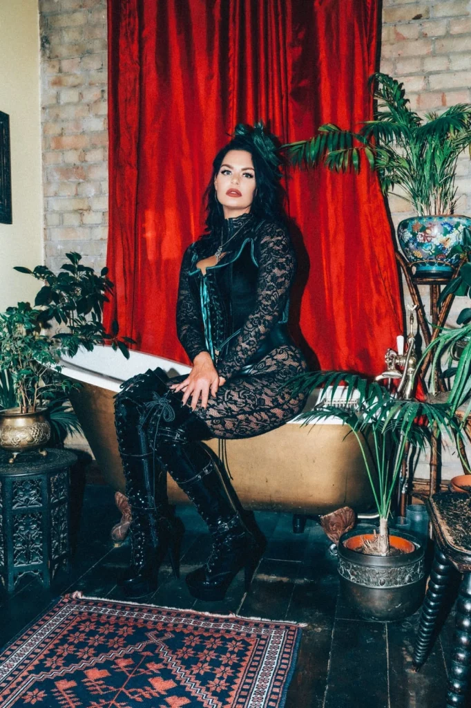 Mistress Charlotte seated on the edge of a bath before a red velvet curtain