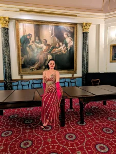 Mistress Charlotte O’Winter in a red gown and pink evening gloves in a grand salon, feminisation mistress in London