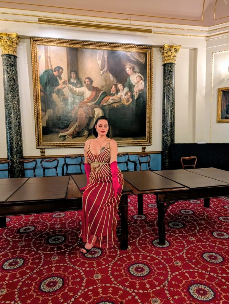 Mistress Charlotte O’Winter in a red gown and pink evening gloves in a grand salon, feminisation mistress in London